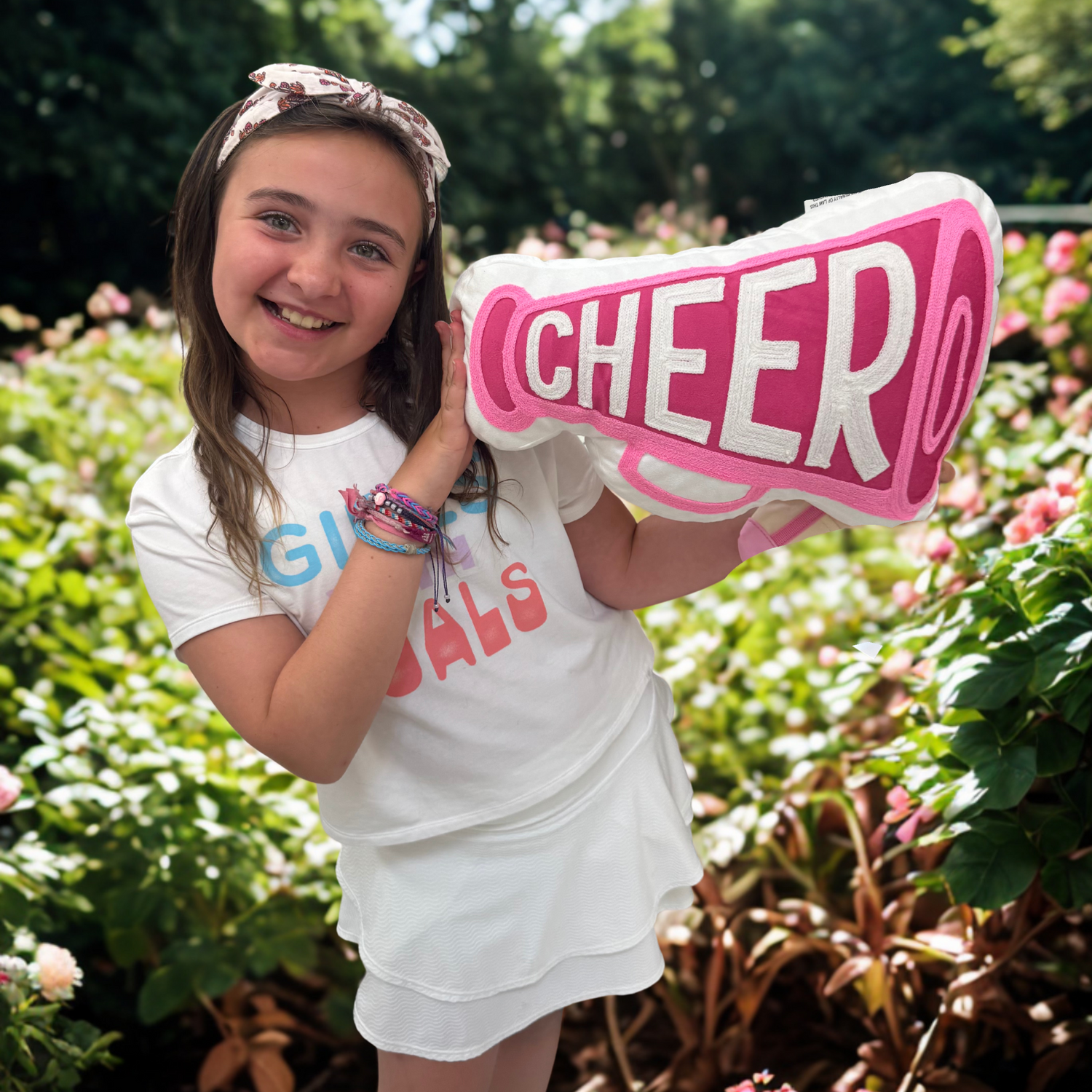 Girl holding a 'CHEER' sign outdoors with greenery and flowers in the background